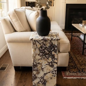 May include: A tall, rectangular pedestal stand crafted from white and dark purple mottled stone with a smooth, polished finish. A matte black vase containing a sprig of dried eucalyptus leaves sits centred on the flat top surface of the pedestal.