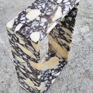 May include: A rectangular marble side table with a unique, open design. The table features a striking pattern of white, cream, and dark brown veining. The overall shape is a continuous loop, creating a modern and artistic aesthetic. The table is made of solid marble.