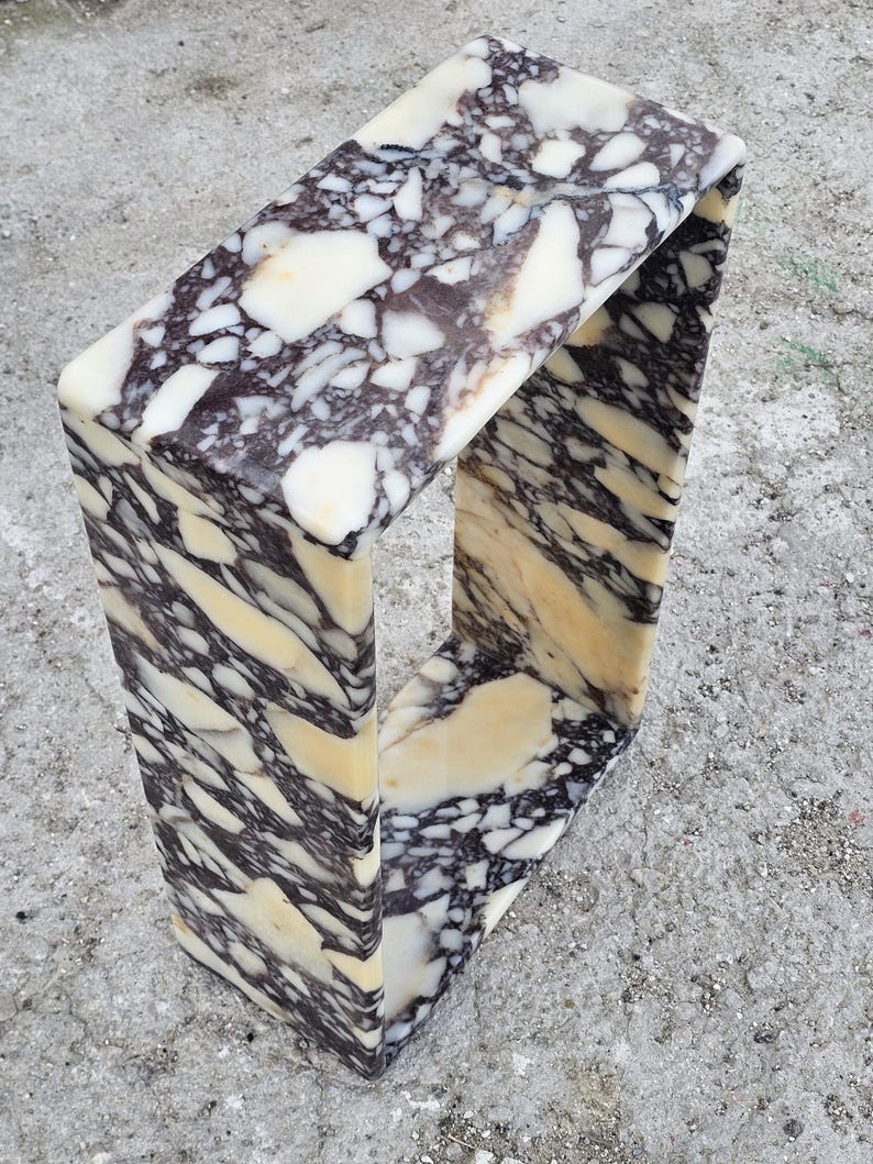 May include: A rectangular marble side table with a unique, open design. The table features a striking pattern of white, cream, and dark brown veining. The overall shape is a continuous loop, creating a modern and artistic aesthetic. The table is made of solid marble.