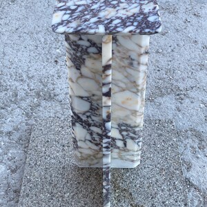 May include: A tall, rectangular pedestal stand crafted from white marble with prominent dark purple and brown veining. The structure features a flat square top surface supported by a central vertical column with a cross-shaped base design.