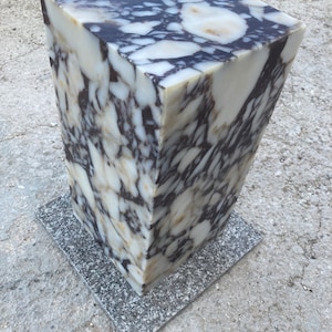May include: A marble pedestal with a rectangular top and a trapezoidal body. The stone features a white base with dark purple and brown veining. It rests on a square, gray granite base. The pedestal is suitable for displaying sculptures or plants.