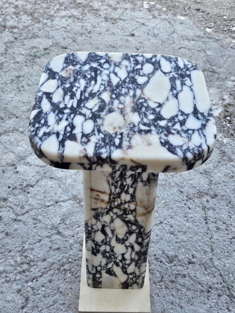 K&ouml;nnte beinhalten: Ein Marmor-Sockeltisch mit rechteckiger Platte und quadratischem Sockel. Der Tisch weist ein auff&auml;lliges Muster aus schwarzen und wei&szlig;en Adern im gesamten Stein auf. Der Sockel ist etwa 76 cm hoch.