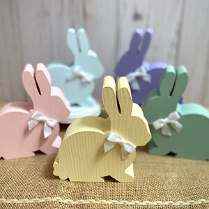 May include: Five wooden bunny figurines in various pastel colors, including pink, light blue, lavender, yellow, and green. Each bunny has a white bow tied around its neck.