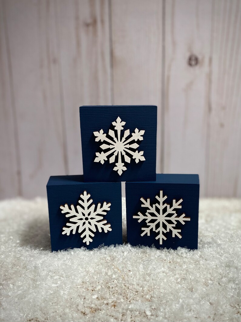 Op de afbeelding: Drie marineblauwe houten blokken met witte sneeuwvlokuitsparingen. De blokken zijn in een driehoekige vorm gerangschikt, met de sneeuwvlokken naar voren gericht. De blokken staan op een bed van witte, kunstmatige sneeuw.