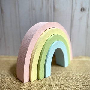 May include: A pastel-colored wooden rainbow toy. The rainbow is made of five arched pieces in shades of pink, yellow, green, and blue. The toy is designed for stacking and play. The rainbow is sitting on a burlap surface.