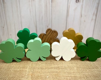 May include: Seven wooden shamrock shapes in various shades of green, brown, and white. The shamrocks are arranged in a row on a burlap surface.