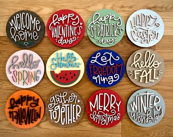 May include: A collection of twelve round wooden signs with various holiday and seasonal greetings. The signs feature phrases like "Welcome Home," "Happy Valentine's Day," and "Hello Spring" in a mix of colors and fonts.