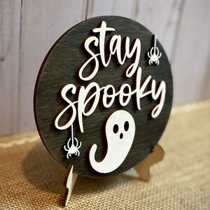 May include: A round wooden sign with a black background and white lettering that reads "stay spooky". The sign has a white ghost and two white spiders hanging from it. The sign is on a wooden stand.