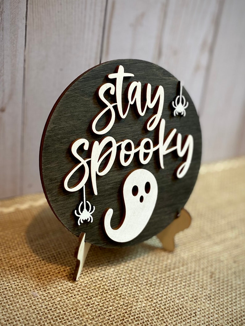 May include: A round wooden sign with a black background and white lettering that reads "stay spooky". The sign has a white ghost and two white spiders hanging from it. The sign is on a wooden stand.