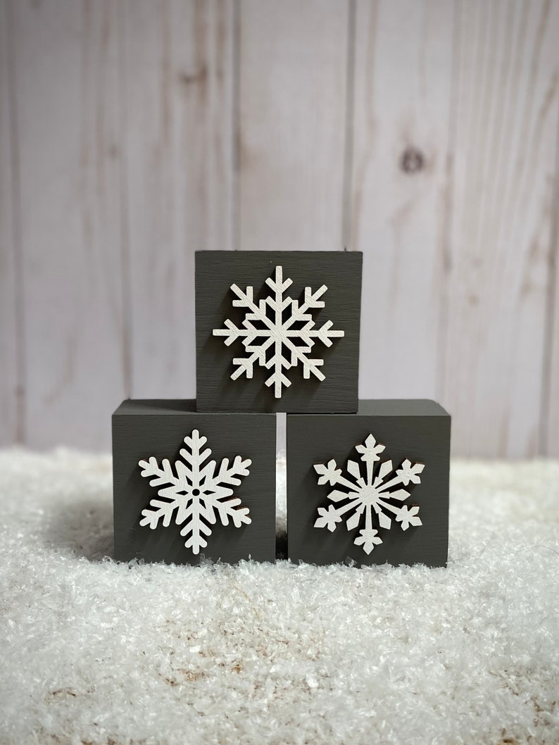 Op de afbeelding: Drie grijze houten blokken met witte sneeuwvlokuitsparingen. De blokken zijn gerangschikt in een kleine piramide en liggen op een bed van wit, zacht materiaal. De achtergrond is een lichtgrijs houten paneel.
