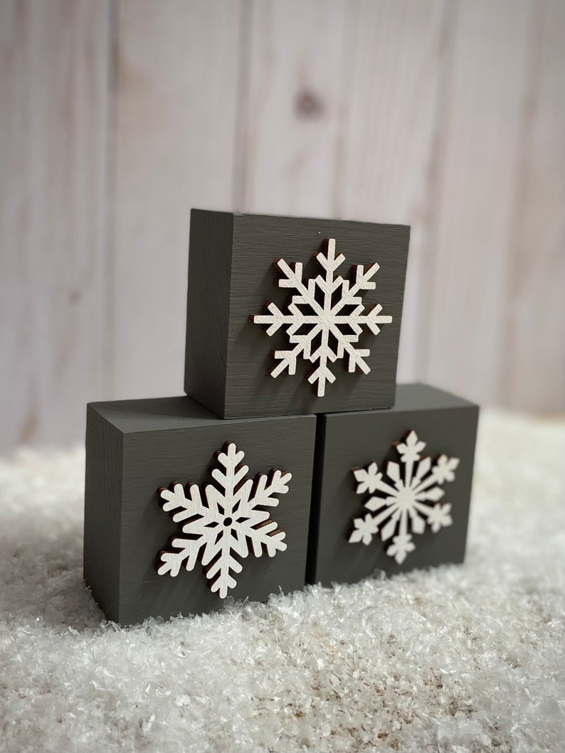 Op de afbeelding: Drie grijze houten blokken met witte sneeuwvlok uitsparingen. De blokken zijn gerangschikt op een bed van kunstsneeuw. De sneeuwvlokken hebben een gedetailleerd, gelaagd ontwerp, ideaal voor winterdecoratie.