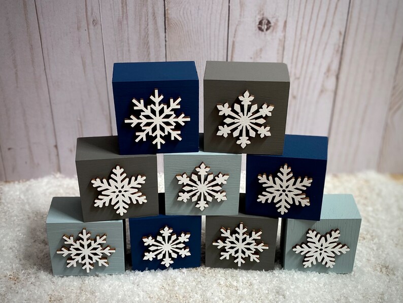 Op de afbeelding: Tien houten blokken in verschillende tinten blauw en grijs, elk versierd met een witte sneeuwvlok. De blokken zijn in een piramidevorm gerangschikt. Perfect voor winterdecoraties.