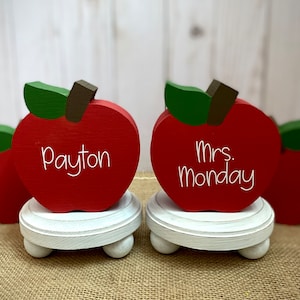 May include: Two red wooden apples with green leaves and white painted names on them. The apples are on white wooden stands with round legs. The names on the apples are "Payton" and "Mrs. Monday".