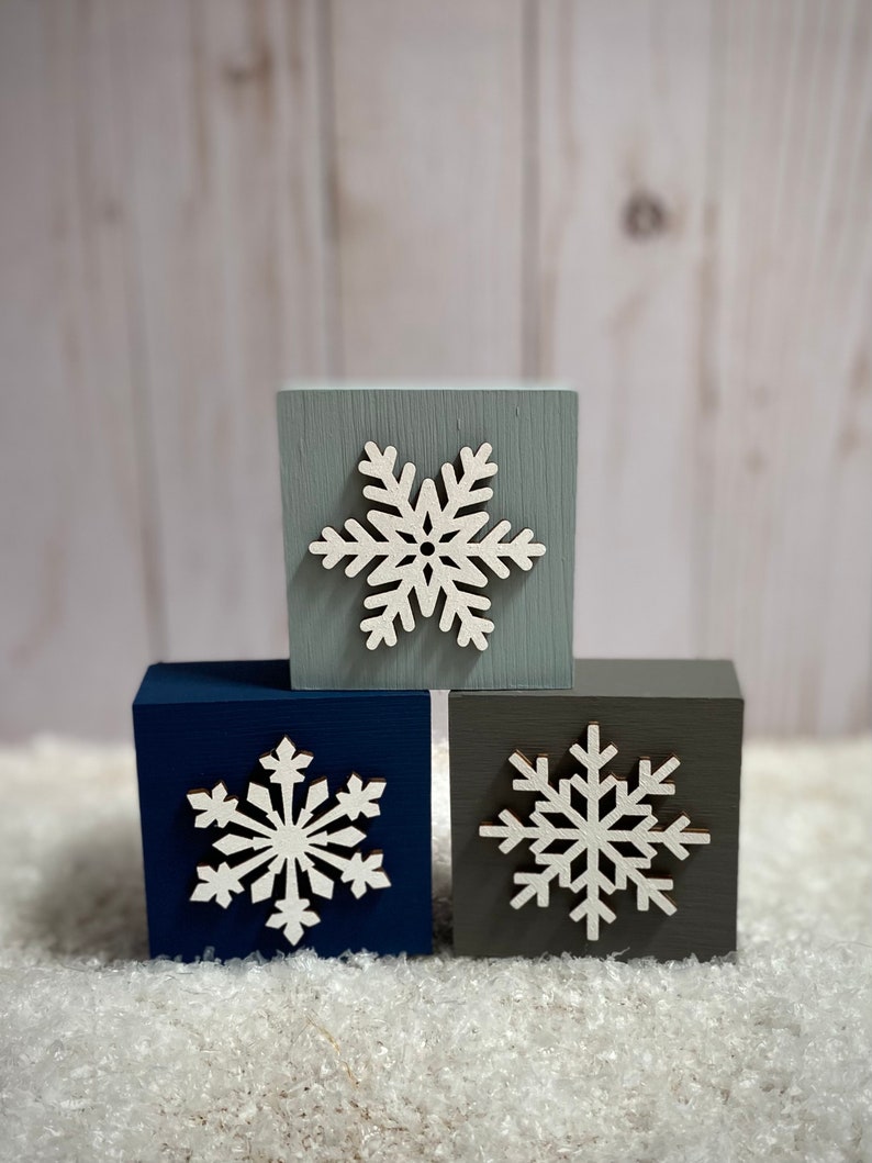 Op de afbeelding: Drie houten blokken in tinten blauw, grijs en marine, elk versierd met een witte sneeuwvlok. De blokken zijn gestapeld en cre&euml;ren een winterse decoratieve display. De achtergrond is een lichte houtnerf.