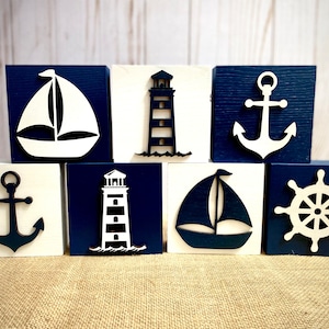 May include: Set of eight wooden blocks with nautical themes. Each block features a different design, including a sailboat, lighthouse, anchor, and ship's wheel. The blocks are painted in shades of blue and white.