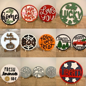 May include: A collection of 12 round wooden signs with various designs and sayings. The signs are painted in different colors, including red, green, blue, black, white, and orange. Some of the sayings include "Love You More", "Love You", "Lucky", "Boo", "Trick or Treat", "Farm Fresh", "Merry Christmas", "Fresh Lemonade 50c", "Let It Snow", "Winter Wishes", "Hey Winter", and "Liberty".