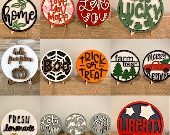 May include: A collection of 12 round wooden signs with various designs and sayings. The signs are painted in different colors, including red, green, blue, black, white, and orange. Some of the sayings include "Love You More", "Love You", "Lucky", "Boo", "Trick or Treat", "Farm Fresh", "Merry Christmas", "Fresh Lemonade 50c", "Let It Snow", "Winter Wishes", "Hey Winter", and "Liberty".