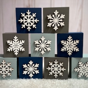 Op de afbeelding: Tien vierkante houten blokken in tinten blauw, grijs en blauwgroen, elk versierd met een witte sneeuwvlok. De blokken zijn in een gestapelde formatie gerangschikt, waardoor een winterse decoratieve weergave ontstaat.