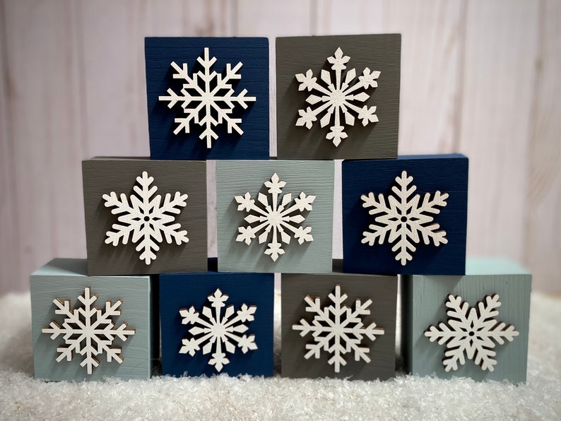 Op de afbeelding: Tien vierkante houten blokken in tinten blauw, grijs en blauwgroen, elk versierd met een witte sneeuwvlok. De blokken zijn in een gestapelde formatie gerangschikt, waardoor een winterse decoratieve weergave ontstaat.