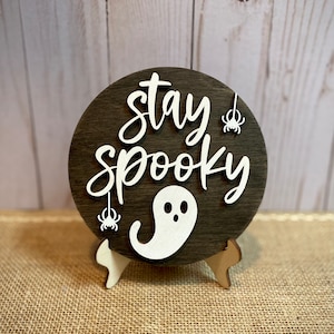 May include: A round wooden sign with the text "Stay Spooky" in white lettering. The sign has a white ghost and two white spiders hanging from it. The sign is on a small wooden stand.