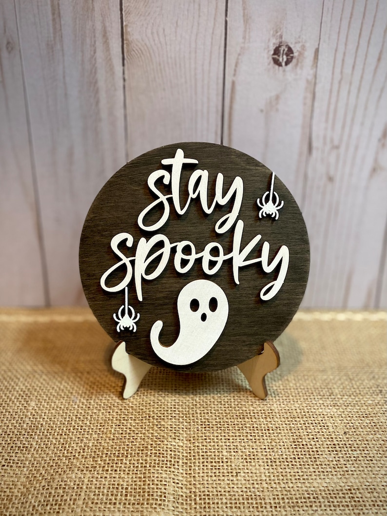 May include: A round wooden sign with the text "Stay Spooky" in white lettering. The sign has a white ghost and two white spiders hanging from it. The sign is on a small wooden stand.