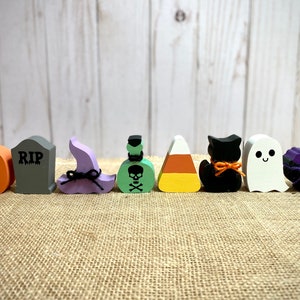 May include: A set of nine Halloween-themed wooden decorations. The decorations include a pumpkin, a tombstone with the word "RIP" on it, a witch's hat, a poison bottle, a candy corn, a black cat, a ghost, and a purple spider web. The decorations are painted in various colors, including orange, green, purple, black, and white.