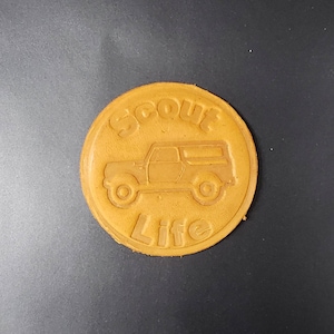 May include: A round, brown leather patch with the words "Scout Life" embossed in a white font. A small, white, cartoon-style jeep is embossed in the center of the patch.
