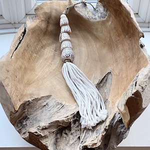 May include: A white tassel with a decorative shell bead accent hangs from a wooden bowl. The bowl is made of natural wood with a rough texture.