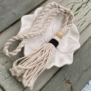 Może przedstawiać: Biały naszyjnik z sznurka makramowego z drewnianym koralikiem i czarnymi koralikami na końcu frędzli. Naszyjnik jest prezentowany na białym ceramicznym talerzu w kształcie kwiatu.