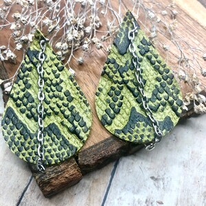 Może przedstawiać: Para zielono-czarnych kolczyków wiszących ze skórą węża z srebrnymi łańcuszkami.