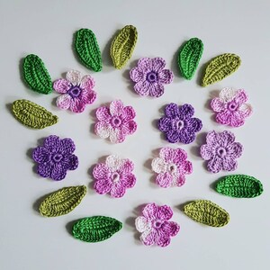 May include: A collection of crocheted flowers and leaves in shades of purple, pink, and green. The flowers have five petals and a centre button. The leaves are long and narrow.