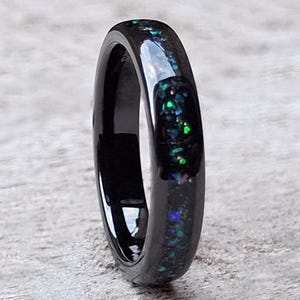 May include: A black ring with a band of iridescent, colorful flecks. The ring has a glossy finish and is set against a light-colored, textured surface. The ring's design is simple and elegant.