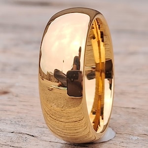 Könnte beinhalten: Ein polierter goldener Ring mit einem abgerundeten, gewölbten Design. Der Ring reflektiert die Umgebung und zeigt eine glatte, glänzende Oberfläche. Der Ring ist ein schlichtes, elegantes Schmuckstück.