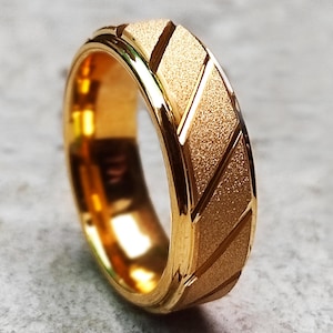 May include: A gold ring with a textured surface and diagonal grooves. The ring has a polished interior and a matte finish on the exterior, creating a contrast in textures. The ring is set against a grey background.