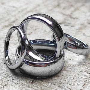 May include: Several polished silver rings of different sizes are arranged together. The rings have a reflective surface, creating highlights. The rings are displayed on a textured, light-coloured wooden surface.