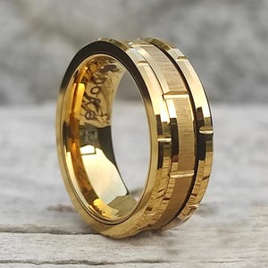 May include: A gold-colored ring with a textured, geometric design. The ring has a polished interior and a matte finish on the exterior, with a series of rectangular and linear patterns. The ring is likely made of a precious metal.