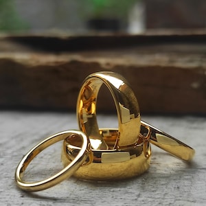 May include: Close-up of several gold wedding rings. The rings are highly polished, reflecting light, and vary in width. The background is blurred, with a wooden surface and greenery visible. The rings are stacked and arranged to showcase their design.