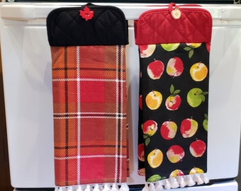 May include: Two hanging kitchen towels with quilted tops and white fringe at the bottom. The left towel features a red, orange, and black plaid pattern with a red maple leaf button. The right towel has a black background with red and yellow apples.
