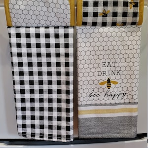 May include: Two kitchen towels hanging on a white stove. One towel features a black and white checkered pattern, while the other has a honeycomb design with the words "EAT DRINK bee happy" and a bee graphic. Both have yellow trim.