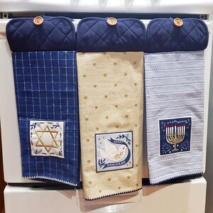 May include: Three kitchen towels with navy blue quilted tops and wooden button closures. The towels feature Hanukkah-themed designs: a Star of David, a dove with "Happy Hanukkah" text, and a menorah.