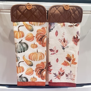 May include: Two hanging kitchen towels with brown quilted tops and wooden buttons. The left towel features a pattern of orange, yellow, and green pumpkins on a white background. The right towel displays a pattern of autumn leaves in shades of red, orange, and brown on a white background.