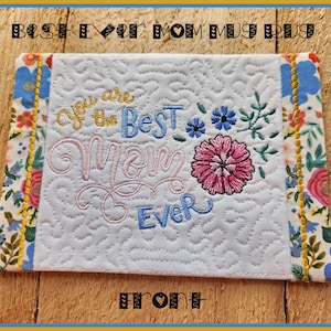 May include: A white quilted mug rug with floral borders and the text "You are the Best Mom Ever" embroidered in pink, blue, and yellow. The rug features a pink flower and blue floral accents. The top reads "Best Ever Mom Mug Rug".