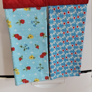 May include: Two hanging kitchen towels with red quilted tops and floral patterned bottoms. One towel has a light blue background with red, yellow, and blue flowers. The other has a blue background with red flowers and white vines. Each towel is held by a decorative flower-shaped clip.
