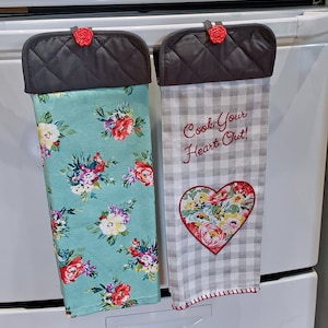 May include: Two kitchen towels hanging on a white oven. One towel is teal with a floral pattern, the other is gray and white checkered with a heart-shaped floral design and the text "Cook Your Heart Out!". Both have gray quilted tops and red rose accents.