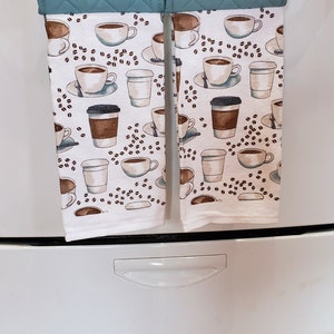 May include: Two teal quilted oven mitts with a white and brown coffee cup pattern hang from a white stove. The mitts are clipped together with a coffee cup-shaped clip. The pattern includes coffee cups, to-go cups, and coffee beans.