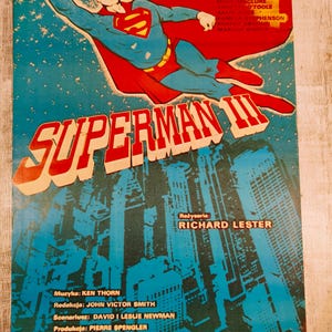 May include: A vintage movie poster for Superman III featuring a blue and red costumed figure flying above a dark blue cityscape. The title Superman III is printed in large red letters. Credits include Christopher Reeve and director Richard Lester.