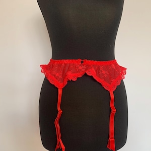May include: A red lace garter belt with adjustable straps and a scalloped edge. The lingerie is displayed on a black mannequin against a white background. The garter belt is a lingerie item.