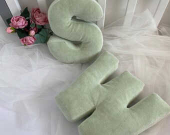 May include: Two large, plush letters, "S" and "W", are made of a soft, light green fabric. The letters are sitting on a white surface with a sheer white fabric draped over them.