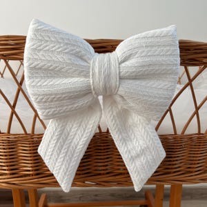 May include: White knitted bow with a cable knit pattern, tied on a wicker crib.