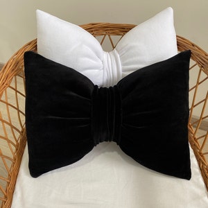 Peut inclure: Deux coussins décoratifs dans un fauteuil en osier. Le coussin du haut est blanc avec une forme de nœud. Le coussin du bas est en velours noir avec une grande forme de nœud.
